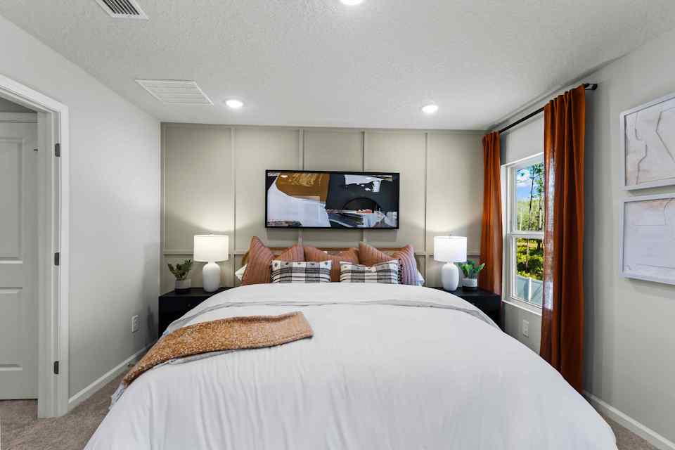 Davor Model Paired Home Primary Bedroom with Orange Bed Comforter and Pillows in Front of Cream Accent Wall at The Landings at Pecan Park