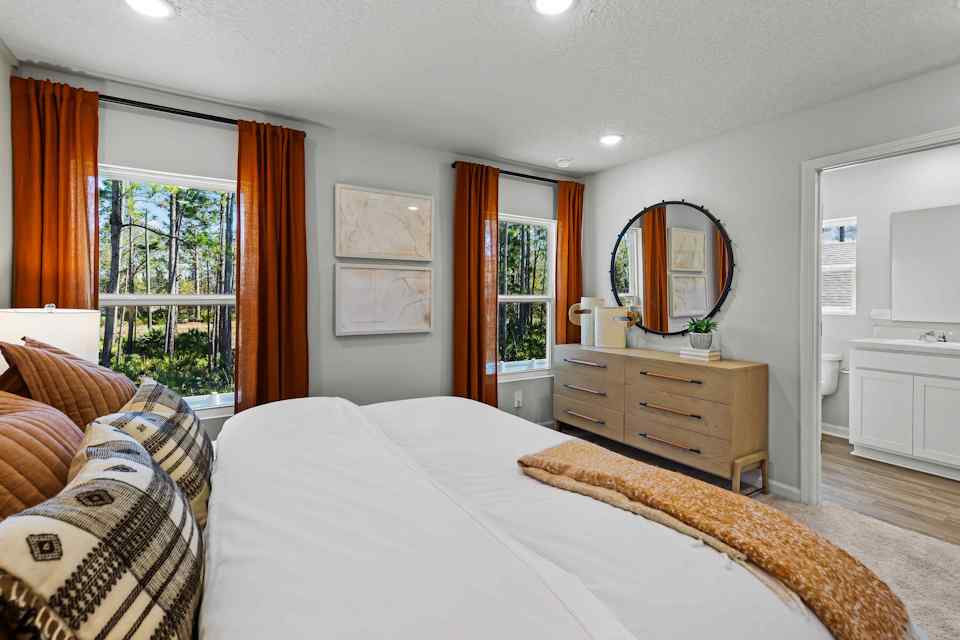 Davor Model Paired Home Primary Bedroom with Orange Bed Comforter and Two Windows with Curtains at The Landings at Pecan Park